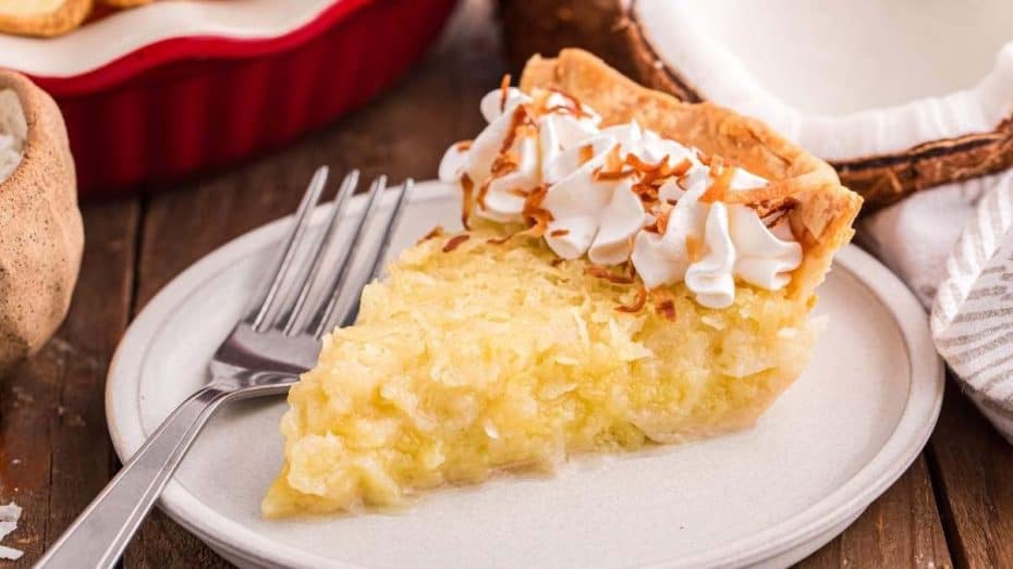 Slice of coconut custard pie topped with whipped cream and toasted coconut flakes on a white plate with a fork beside it.