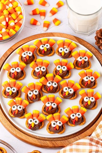 A plate of turkey-shaped treats made with pretzels, candy corn, and chocolate, surrounded by more candy corn and a glass of milk.
