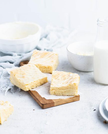 Three white chocolate brownies on a small, rectangular, wooden cutting board with a white bowl, a bottle of milk and a small bowl of white chocolate in the background.