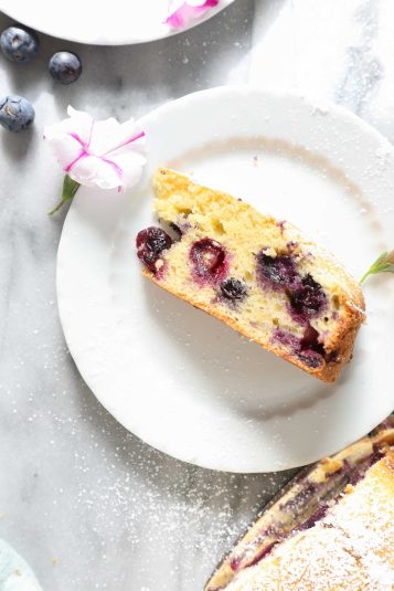A slice of Lemon Blueberry Olive Oil Cake on a small white plate with a light marble background surrounded by powdered sugar, extra blueberries, and purple and white flowers.