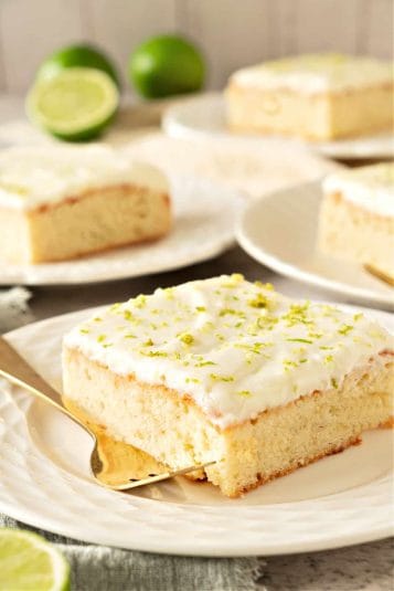 White plates with cream topped cake squares with lime zest. Forks, limes in the gray background.