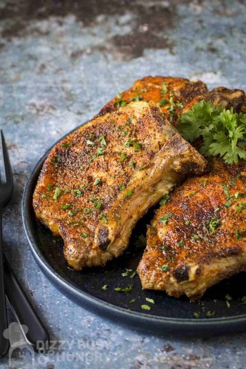 Side shot of three pork chops on a black plate garnished with fresh parsley with a black fork on the side.