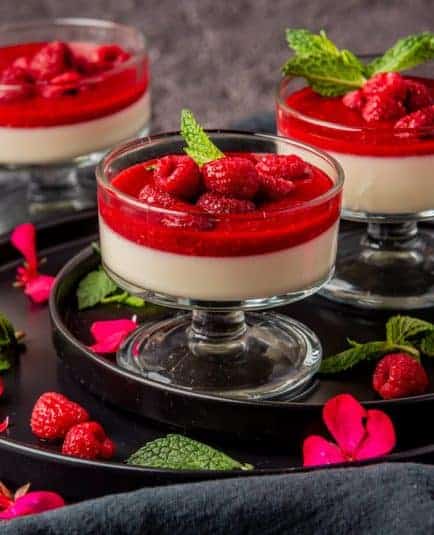 Raspberry panna cotta in glass dishes on black plates. Topped with raspberries and mint leaves, 3 dessert dishes.