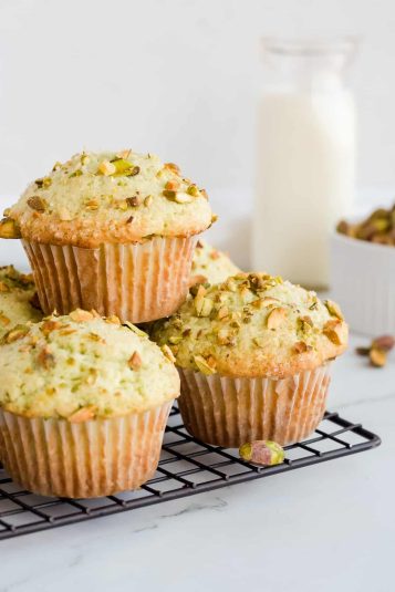 Pistachio muffins stacked on wire rack with a glass of milk and a bowl of pistachios in background.