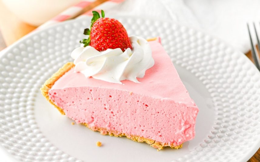 A slice of strawberry pie sits on a white plate. The pink filling and graham cracker crust make it irresistible. Topped with whipped cream and a fresh strawberry.