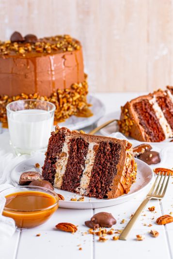 Slices of Turtle Cake shown on a white tile table top with weathered light wood wall, glass of milk and caramel sauce in a bowl.