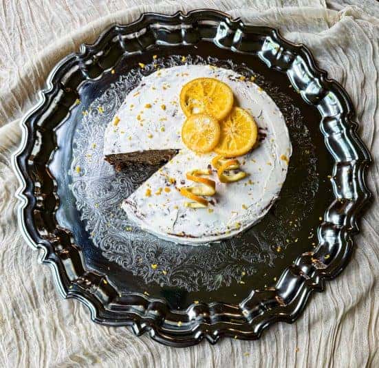 Frosted cake with one piece missing on a silver plate with oranges on top.