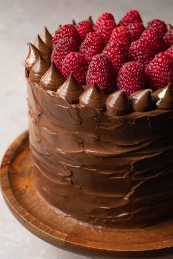 Close up shot of a rustic-looking chocolate cake on a wooden plate covered with silky chocolate cream and decorated with fresh raspberries.
