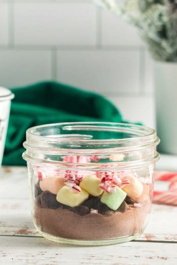 Side view of a short mason jar layered with cocoa powder, chocoalte chips, marshmallows and crushed candy canes.