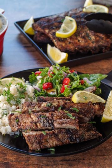 Cut Greek ribs on a plate with Greek rice and salad, with tray of ribs in the background.