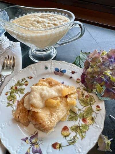 A floral porcelain plate with a scoop of bread pudding drizzled with sauce next to an etched glass pitcher of sauce.