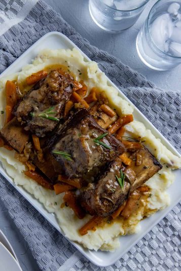 Cooked beef ribs on a bed of mashed potatoes with onions and carrots scattered around.
