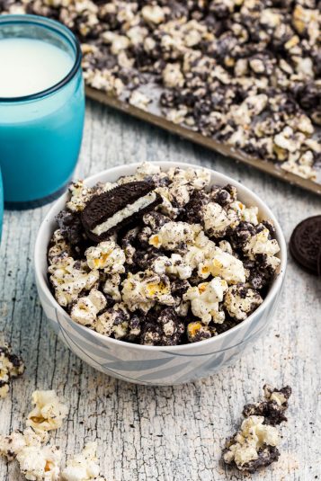Popcorn in a light bowl with crushed Oreo cookies mixed in with white chocolate in front of blue glasses filled with milk.