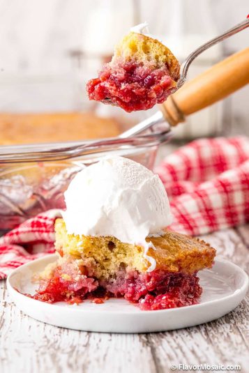 A side view of a white dessert plate with a single serving of raspberry cobbler on it with vanilla ice cream on top that has had a bite taken out of it, and a spoon with a bite of the raspberry cobbler is held above the rest of the serving. In the background is the rest of the cobbler (blurred) in a glass baking dish with a red and white checkered napkin to the right of the dish.