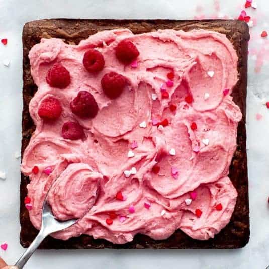 A spoon spreading frosting on raspberry chocolate brownies with fresh raspberries and sprinkles.
