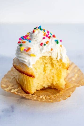 Cream-filled cupcake with colorful sprinkles and vanilla frosting, on a white marble surface.