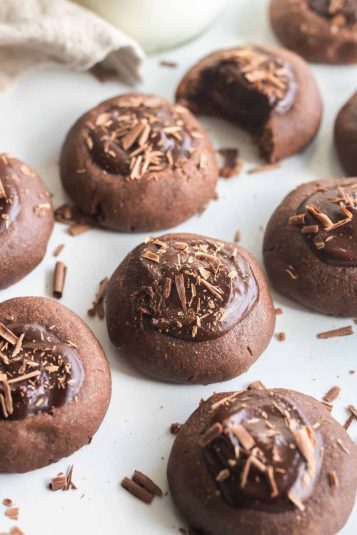 Many raspberry chocolate thumbprint cookies on a staged countertop showing chocolate shavings sprinkled on top of each one.
