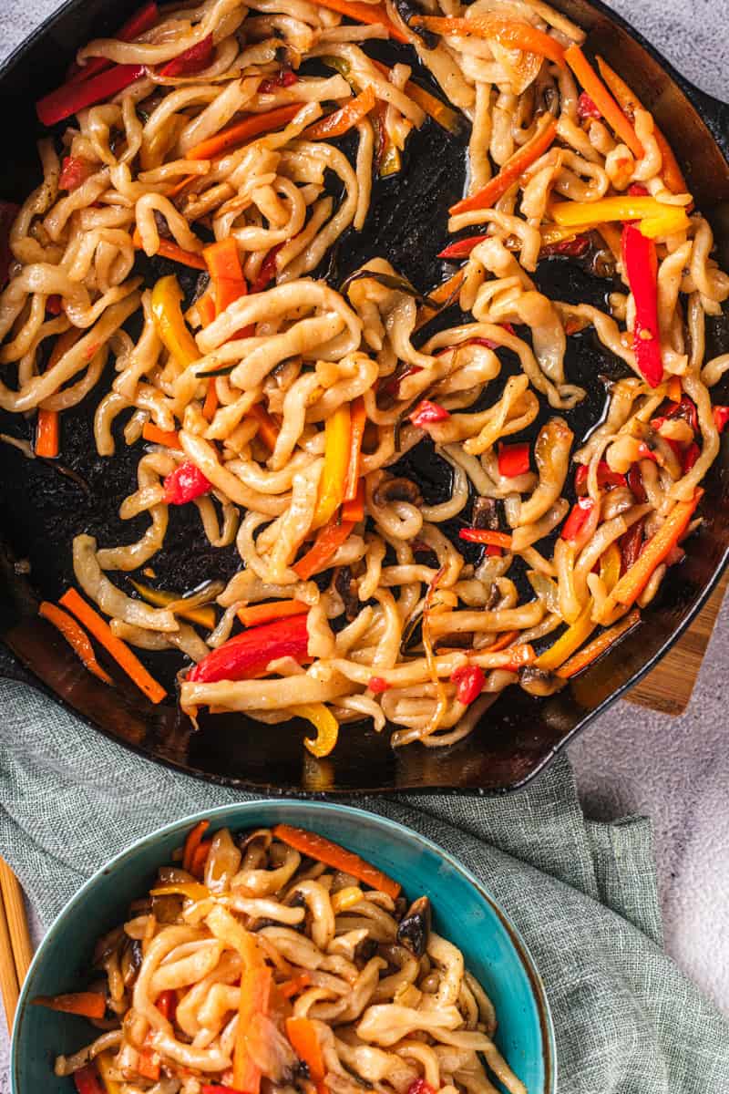 a skillet of stir fried udon noodles with bell pepper, mushrooms, and a spicy sauce.