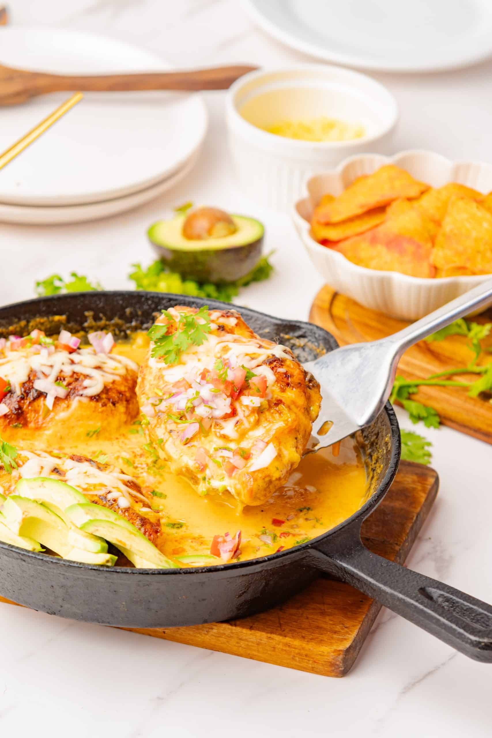 a metal spatula lifting our a chicken breast from queso sauce in a cast iron skillet. 
