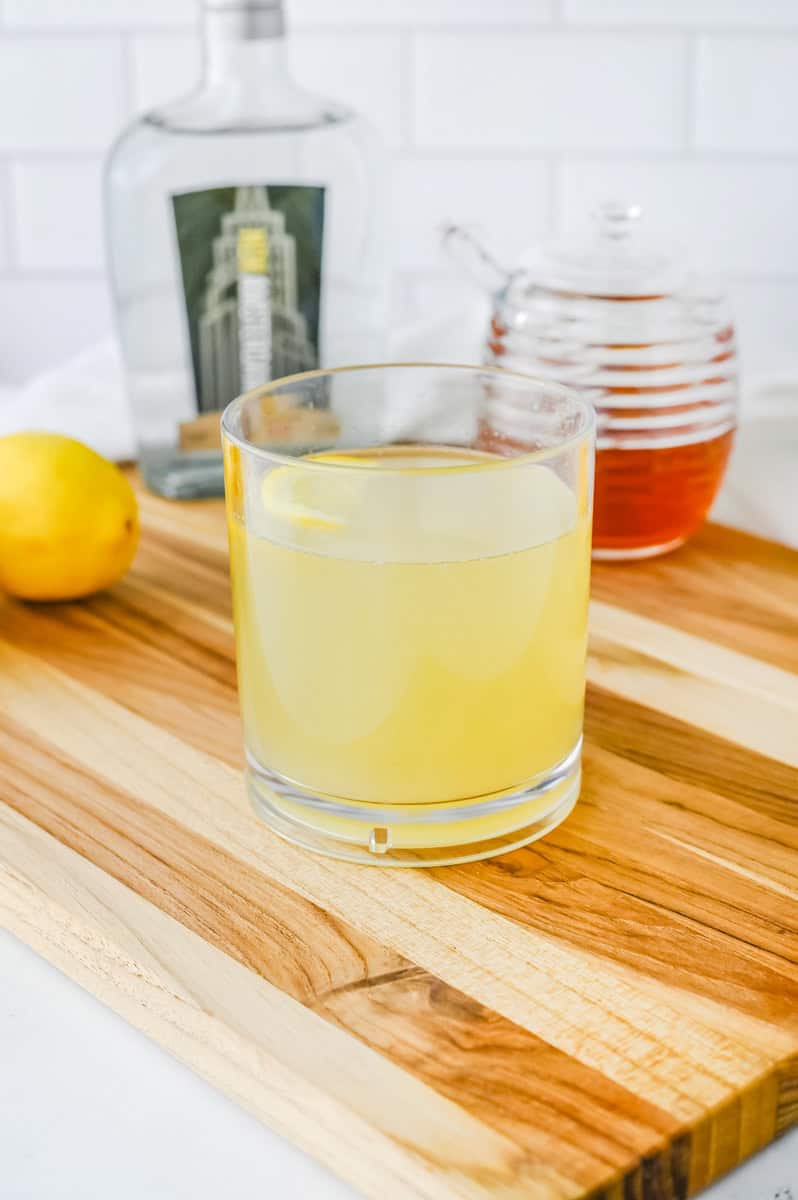 a yellow bee's knees in a rocks glass with gin, honey, and lemon in the background.