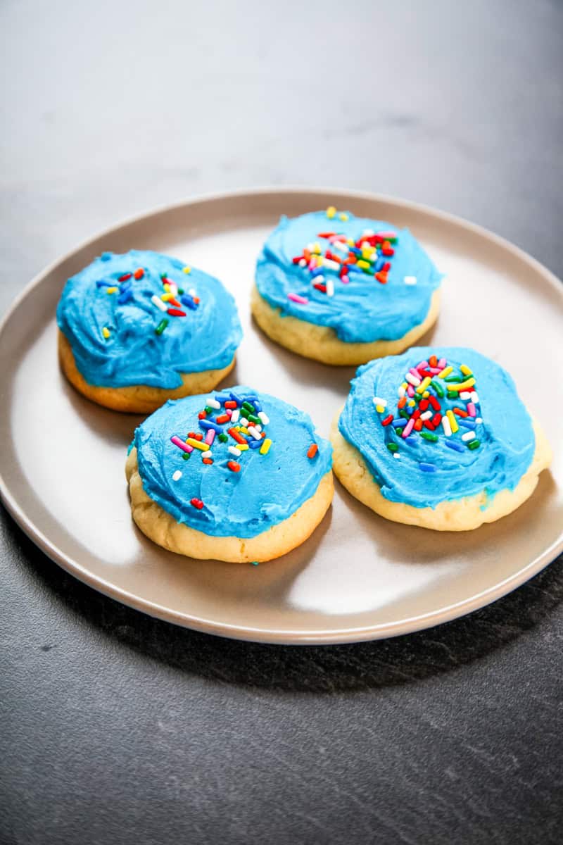 a plate of 4 Lofthouse style cookies with blue icing and rainbow sprinkles.