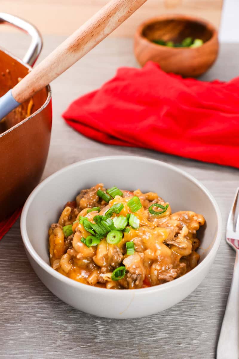 a white bowl of one pot taco pasta topped with green onions on a table with a red dish towel.