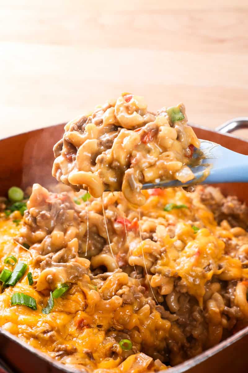 a blue spoon lifting out cheesy taco pasta with ground beef, tomatoes, and green onions.