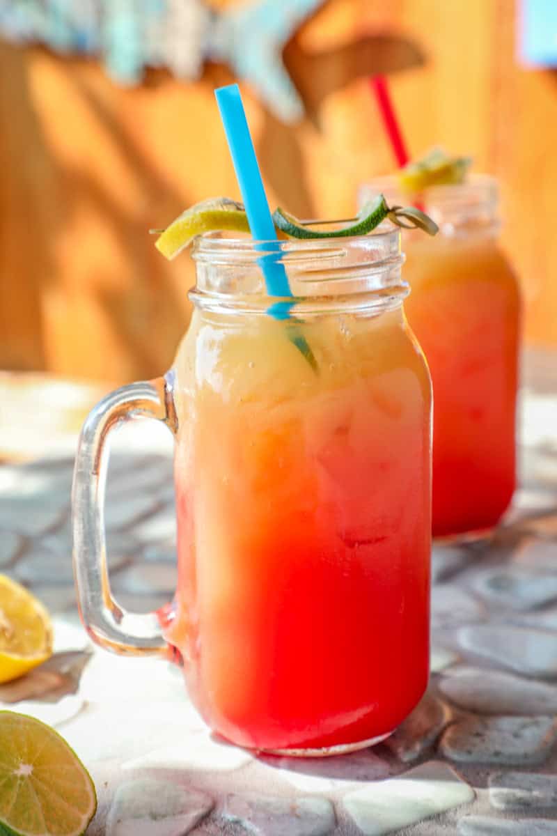 pineapple rum punch in a glass with a handle with a blue straw and a lemon and lime garnish.