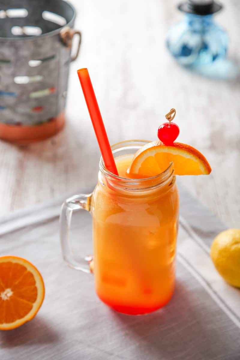 Planters punch cocktail in a glass mug with a straw, orange slice, and maraschino cherry. 