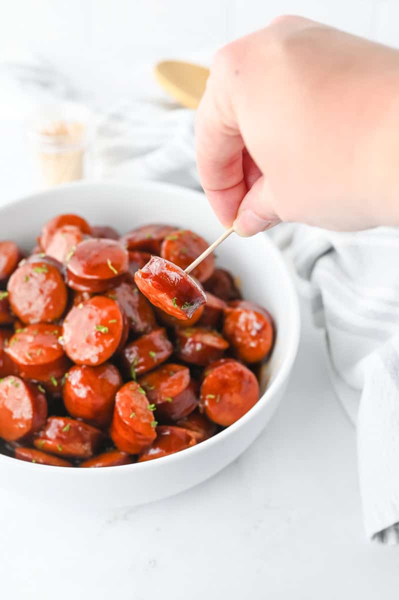 a BBQ kielbasa bite on a toothpick over more BBQ kielbasa in a white bowl.