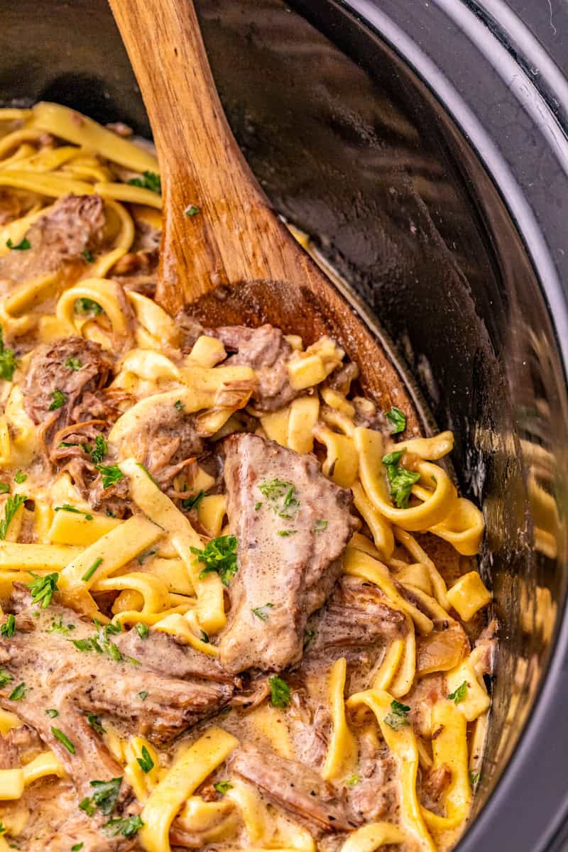 a wooden spoon lifting out crock pot beef and egg noodles in a creamy gravy.