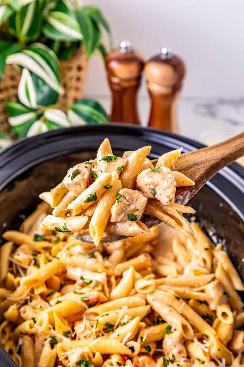 a wooden spoon with cajun chicken and penne pasta over top of a black crock pot.