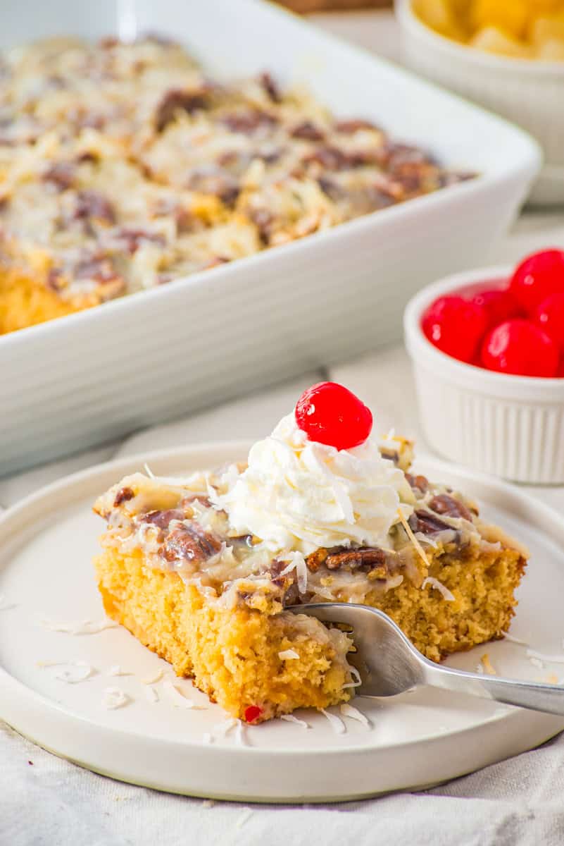 a fork cutting into fruit cocktail cake topped with a coconut pecan topping, whipped cream, and a cherry. 