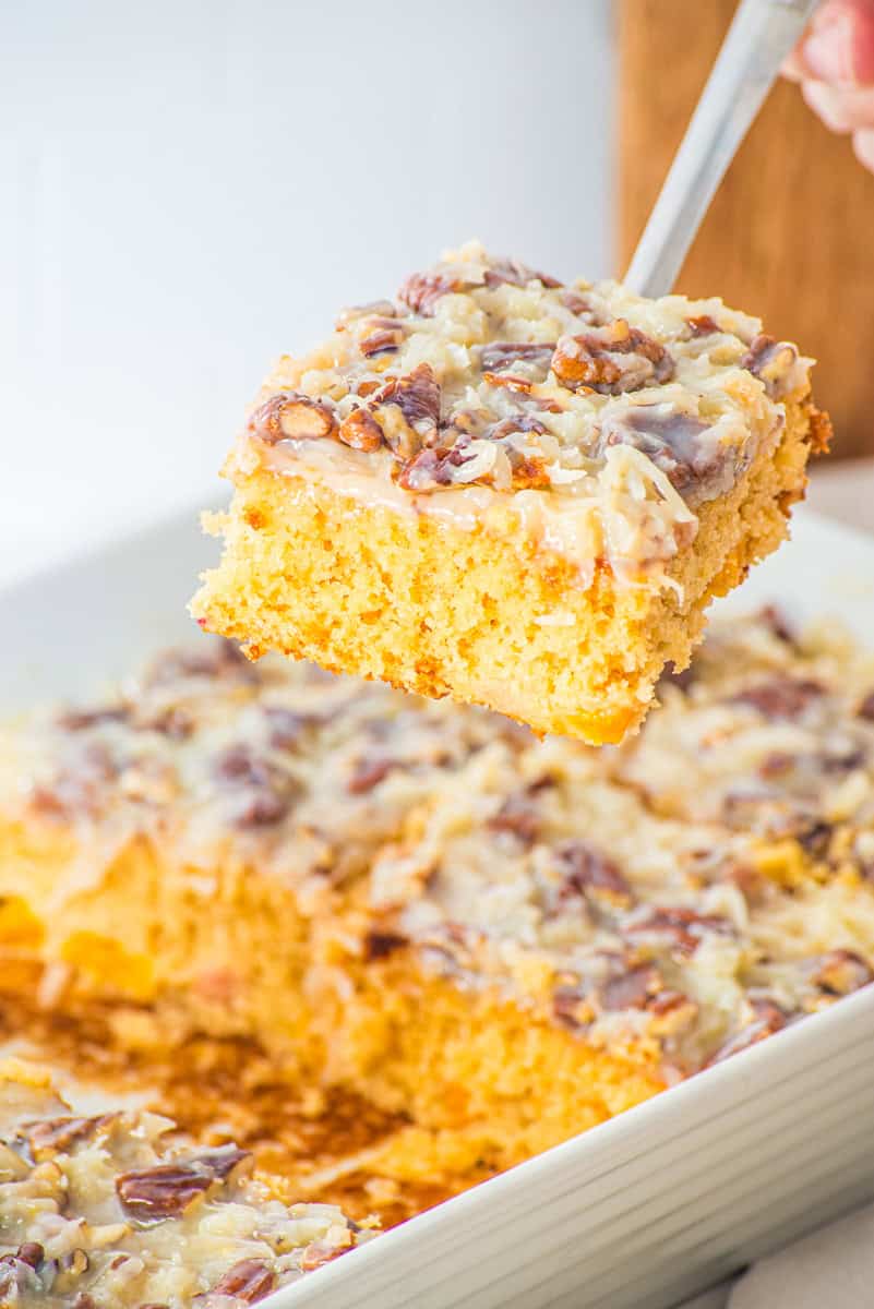 a spatula lifting a slice of fruit cocktail cake with a pecan and coconut topping out of a 9x13 pan.