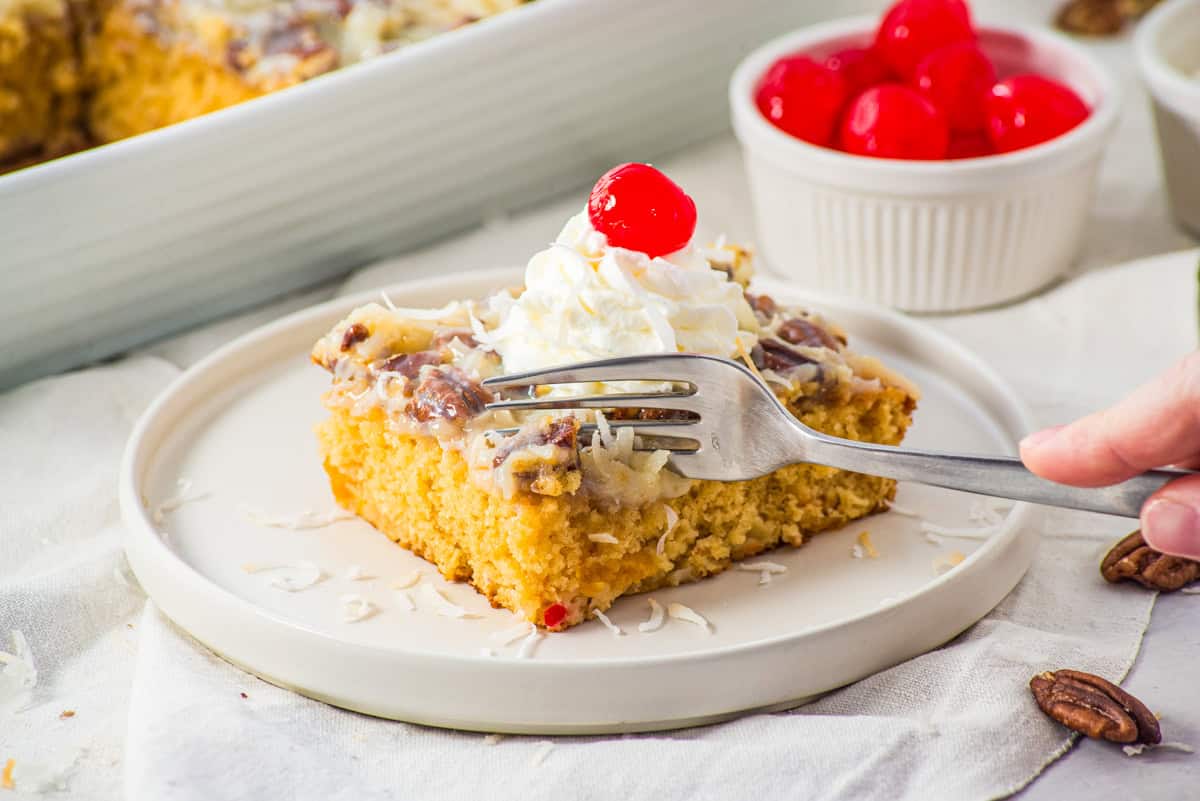 a fork cutting into fruit cocktail cake topped with whipped cream and a cherry. 