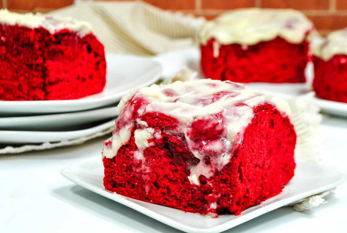 a red velvet cinnamon roll topped with cream cheese frosting on a white plate. 