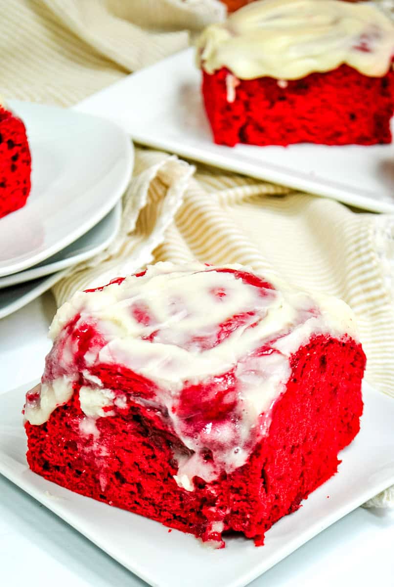 a red velvet cinnamon roll topped with cream cheese frosting on a white plate. 