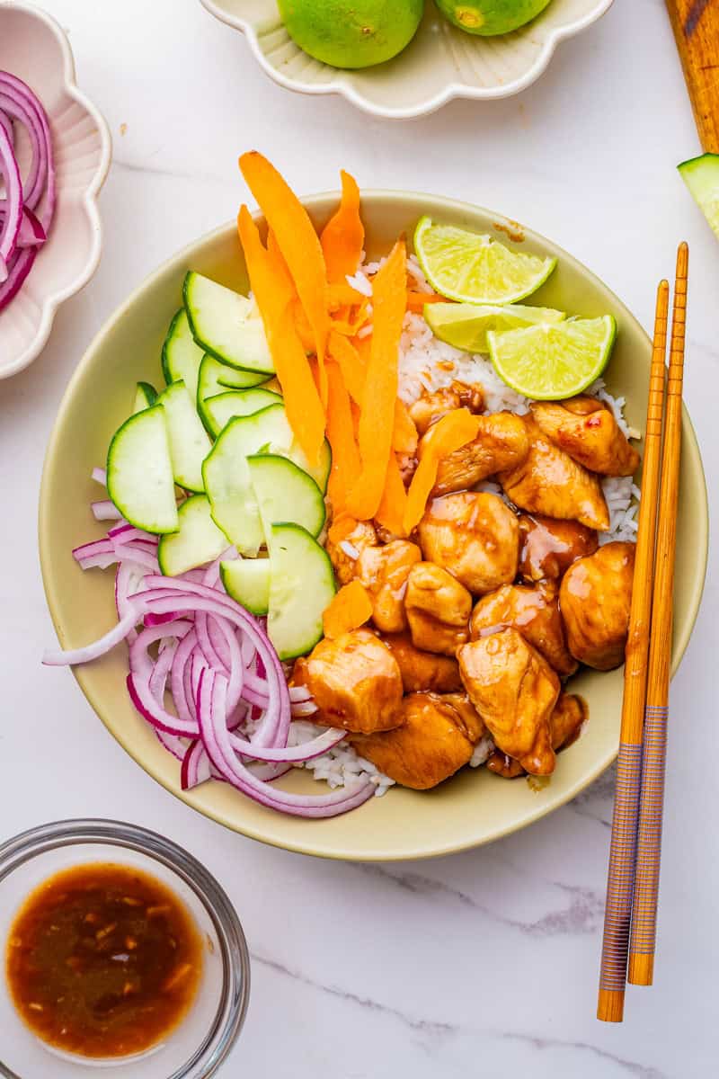 a chicken poke bowl with teriyaki chicken, cucumbers, carrots, red onion, lime slices, and white rice.