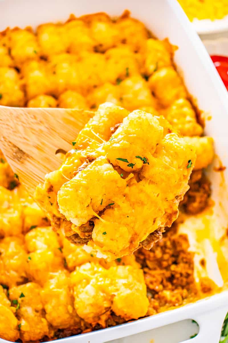 a wooden spoon lifting out a serving of sloppy joe tater tot casserole topped with cheese and parsley.