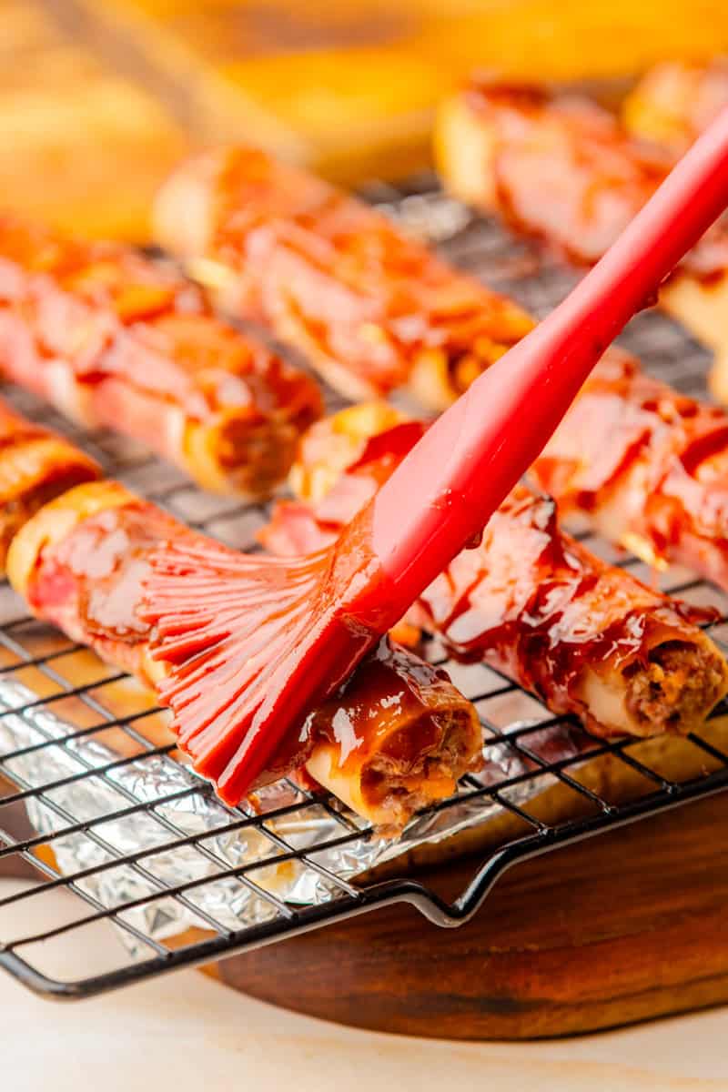 shotgun shells filled with beef and cheese and wrapped in bacon being brushed with BBQ sauce with a silicone brush.