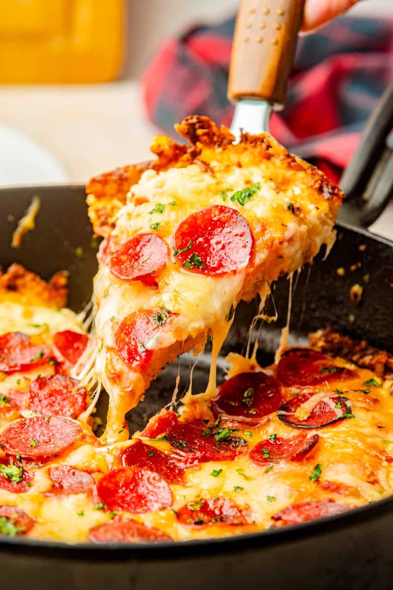 a spatula lifting out a slice of cheese and pepperoni pan pizza from a cast iron skillet.
