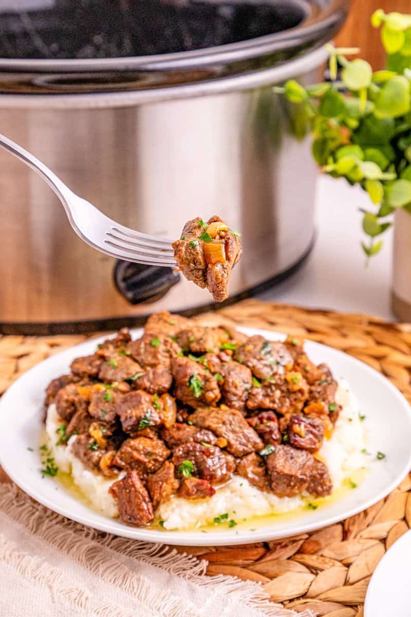 a fork with garlic butter steak bites it onions on it with a plate of steak bites on mashed potatoes below it.