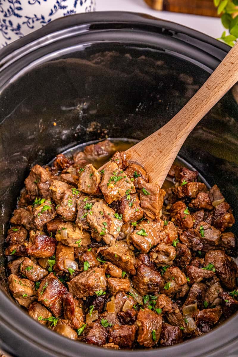 a wooden spoon lifting sirloin steak bites in a ranch butter sauce out of a slow cooker.