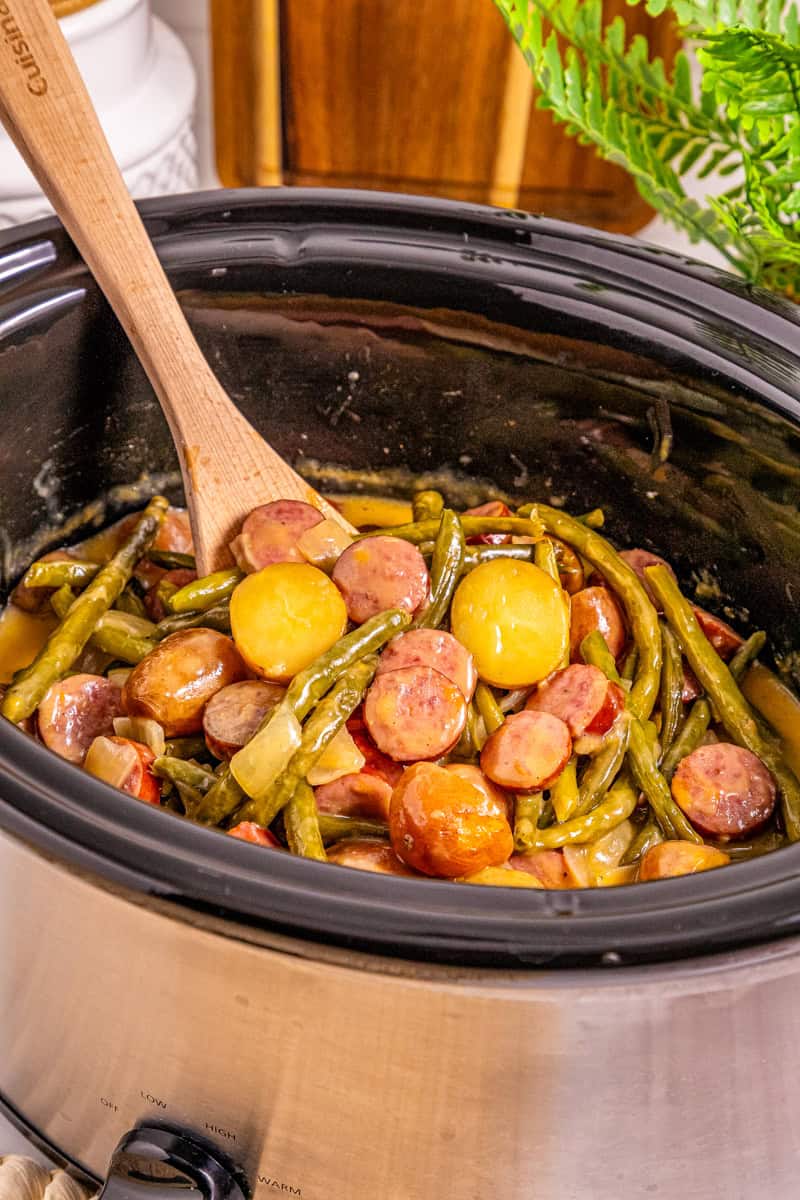a spoon lifting swamp potatoes with sausage and green beans out of the crock pot.