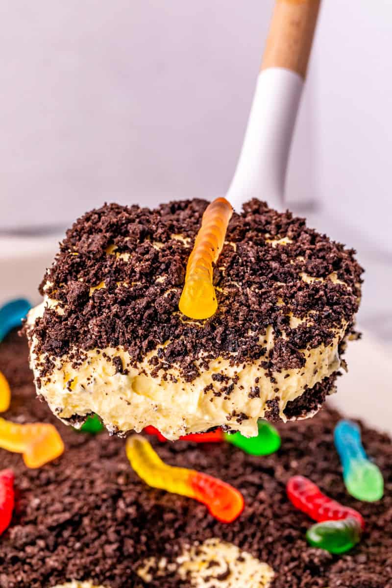 a spatula holding a slice of dirt cake with an oreo crust and topped with a yellow gummy worm.
