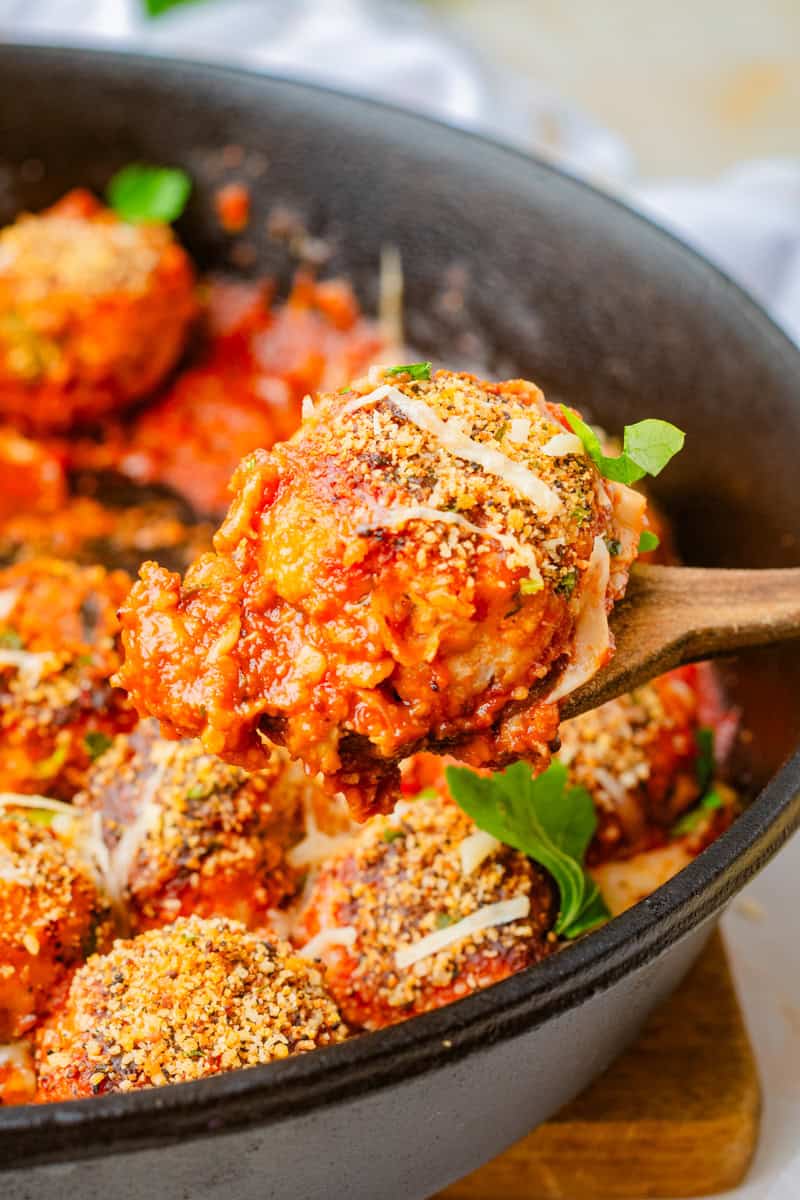 a wooden spoon lifting out a chicken parmesan meatball covered in marinara sauce and topped with a crispy breadcrumb topping.