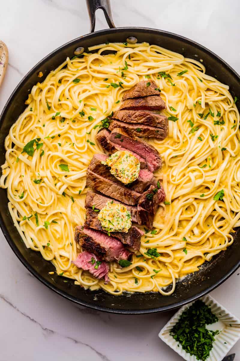 a skillet of fettucine in a cream sauce topped with pan seared ribeye steak topped with cowboy butter.