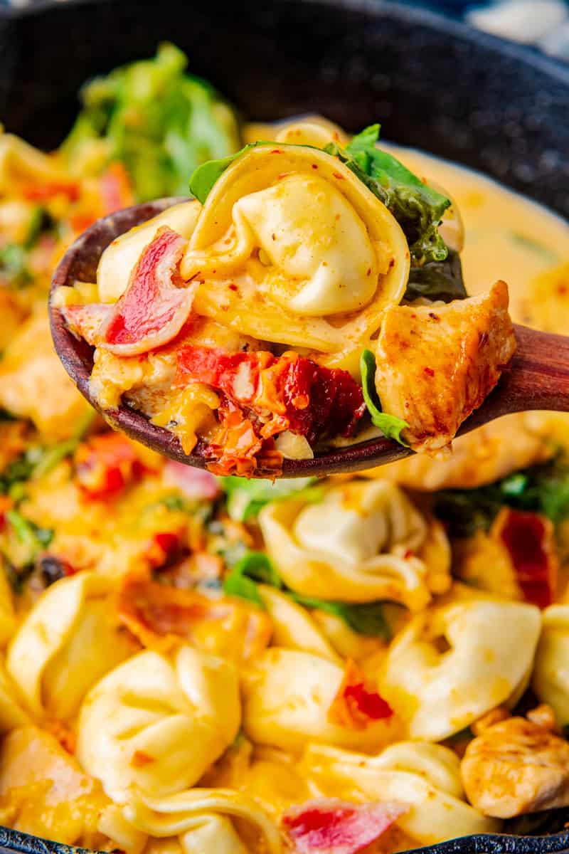a wooden spoon with tortellini, bacon, chicken and spinach in a sun dried tomato cream sauce.