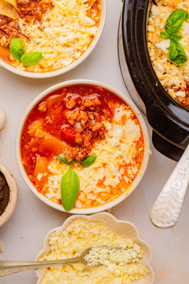 an overhead view of a bowl of lasagna soup topped with cheese with a crock pot in the background.